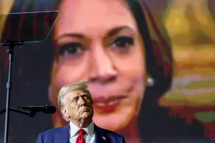 Donald Trump spricht vor seinen Anhängern, während im Hintergrund ein Video über Kamala Harris läuft. 